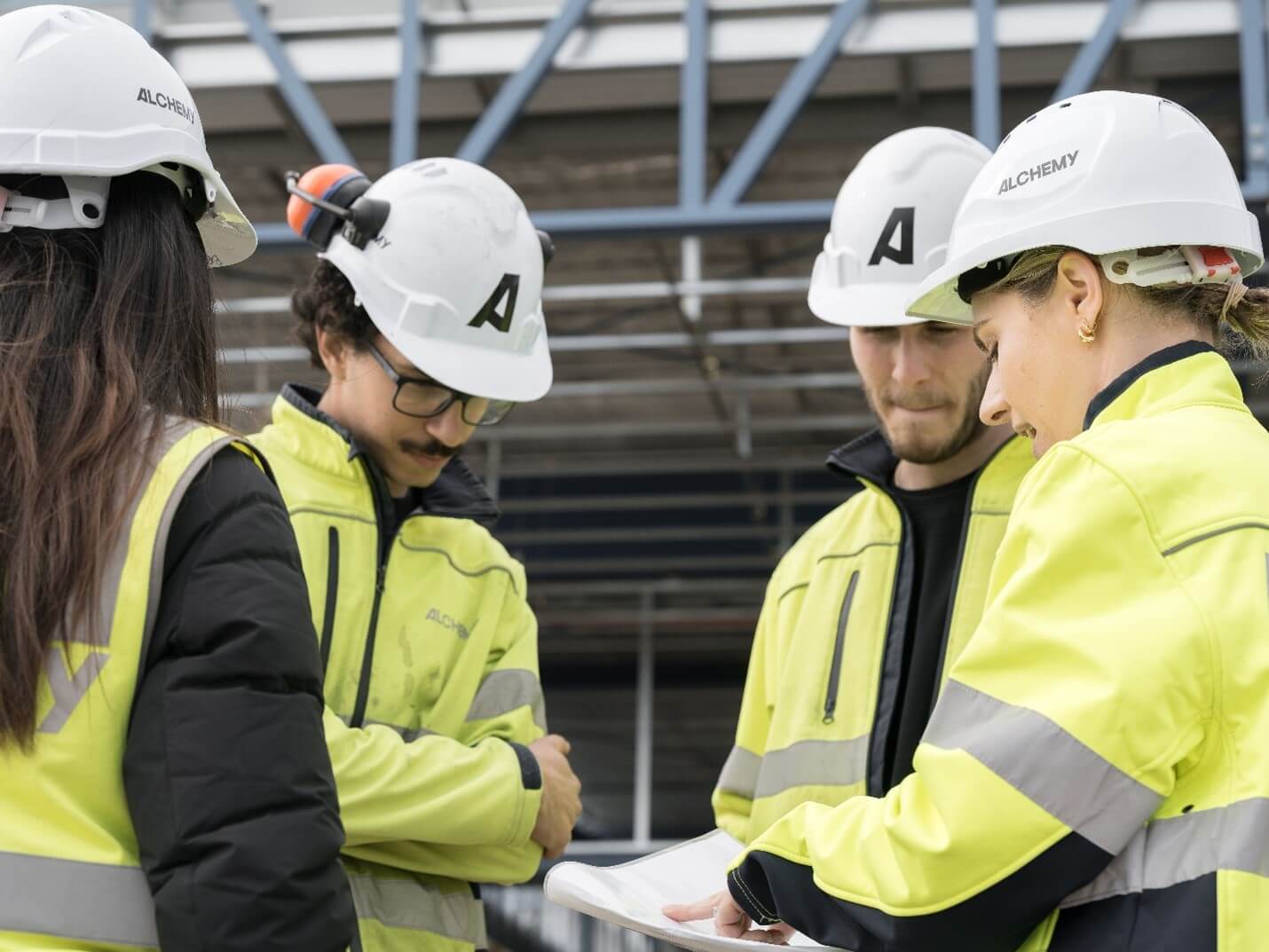 An image of 2 females and 2 males at work in white hard hats with yellow hi-vis standing amongst each other at work, mid discussion, female at front right of the image is pointing to a document while speaking. This image suggests a small team meeting.