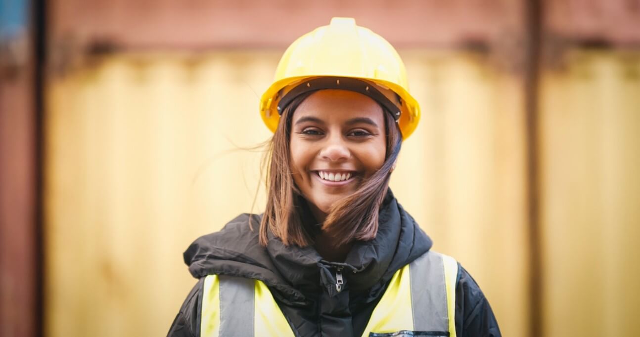 Young woman with medium skin tone and brown hair smiling directly at camera with yellow hard hat, black puffer jacket with hi-vis vest on, her background is stacked shipping containers, suggesting she may work on a port.